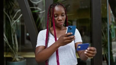 Güzel afro-amerikan kadın, örgülü, akıllı telefonuyla kredi kartının bir fotoğrafını çekiyor, güneşli bir caddede rahatça duruyor.