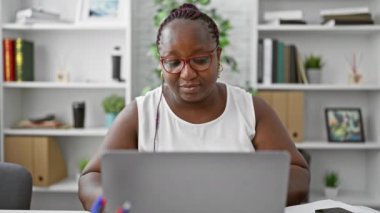 Yorgun, örgü ve gözlüklü Afro-Amerikalı iş kadını, iş yüzünden stresli, ev içi ofiste dizüstü bilgisayarına ara veriyor.