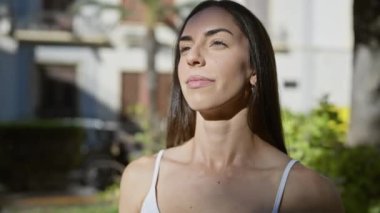 Serene genç İspanyol kadın, gözleri kapalı, parkın yeşilliğinin ortasında dua etmeye dalmış, dışarıdaki sakinlik inancını dengeliyor. Doğadaki rahatlatıcı meditasyonun güzel portresi..