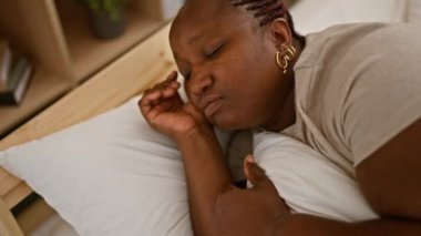 Yorgun Afrikalı Amerikalı kadın rahat yatak odasında dinleniyor, rahat yatağında uyuyor, yumuşak bir battaniyenin altında yorulmadan dinleniyor, yastığın üzerine örülmüş saçlar serpiştiriyor.