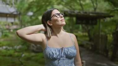 Güzel İspanyol kadın, gözleri kapalı, nefes nefese, Japon yeşil parkında duruyor, güneş ışığının tadını çıkarıyor, gözlük takarak meditasyon yapıyor.