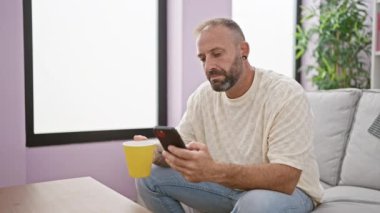 Şaşırmış genç bir adam, evinde koltukta oturmuş, sabah espresso içiyor, akıllı telefon ekranına şaşkın bir ifadeyle bakıyor.