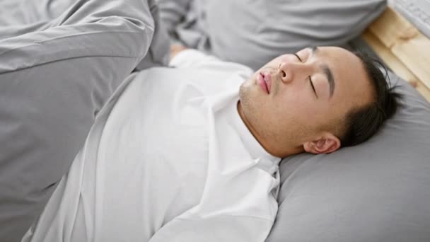居心地の良いパジャマで疲れた若い中国人男性は 家の親密なベッドルームのインテリアを通して座っている暖かい朝の光の最中にプッシュベッドに横たわって 至福の眠りに甘い快適さを見つける — ストック動画