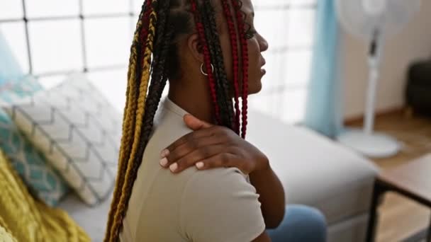 美しいアフリカ系アメリカ人女性は リビングルームのソファーに座ってストレスと傷つき 背中に触れ 心配した表情で自宅のバッカチに苦しんでいます — ストック動画