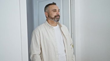 Sakallı, kır saçlı, olgun İspanyol bir adam. Eve gelen misafirleri karşılıyor..