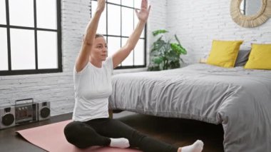 Parlak sarışın kadın meditasyon yapıyor, sabah yoga rutini için yatak odasında bacaklarını açıyor.