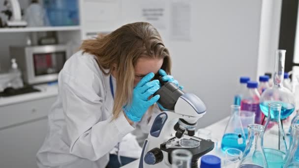 一位严肃而专注的金发女科学家 专心致志地从事重要的医学研究 使用显微镜 并在繁忙的实验室里做笔记 — 图库视频影像