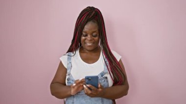 Kendine güvenen Afro-Amerikalı kadın akıllı telefondan sevinçle mesaj atıyor, pembe arka planda gülümsüyor.