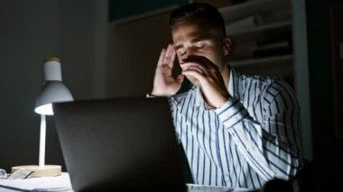 Başı ağrıyan yorgun genç adam, loş ofiste dizüstü bilgisayarla geç saatlere kadar çalışıyor..
