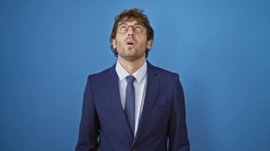 Şaşkın bir genç adam iş elbisesi içinde, şaşırmış bir şekilde tek başına mavi arka planda, şaşkın bakışlı ve neşeli bir jest yapıyor.