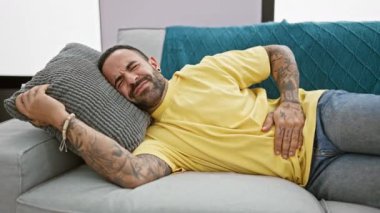 Sarı gömlekli sakallı bir adam, karnını tutarken acı içinde kıvranıyor, gri bir kanepede uzanıyor..