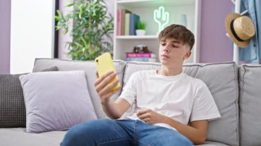 Genç bir adam, rahat bir oturma odasında gri bir kanepede otururken telefonuyla selfie çekiyor..