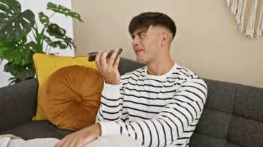 Mutlu İspanyol genç adam, akıllı telefonundan güvenli bir şekilde sesli mesaj gönderiyor. Oturma odasındaki kanepede rahatça oturuyor.