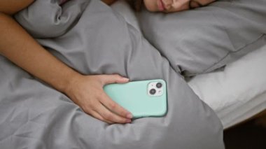 Uykulu genç İspanyol kadın, rahat bir şekilde yatakta uzanıyor, rahat bir yatak odasında akıllı telefonunu tutuyor - yorgun bir günün ardından evde dinlenen çekici bir kadının portresi