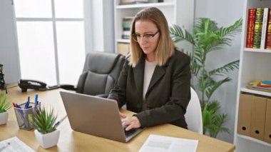 Kendine güvenen ve güzel sarışın iş kadını neşeli bir şekilde laptopuyla canlı ve zarif bir ofis ortamında çalışıyor.