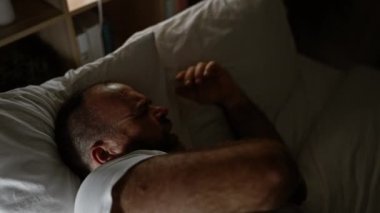 Yatakta yatan yakışıklı beyaz adam yorgun, karanlık yatak odasında uykusuzluk çekiyor, evin arka planı, stresli bir gecenin ortasında hafifçe başucu lambasıyla aydınlatılıyor.