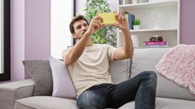 İspanyol adam, oturma odasının iç tarafındaki kanepede akıllı telefonuyla selfie çekiyor.