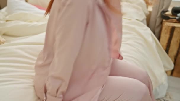 居心地の良い寝室でリラックスしたピンクのパジャマの若い女性 レジャー感と快適さを伝える — ストック動画