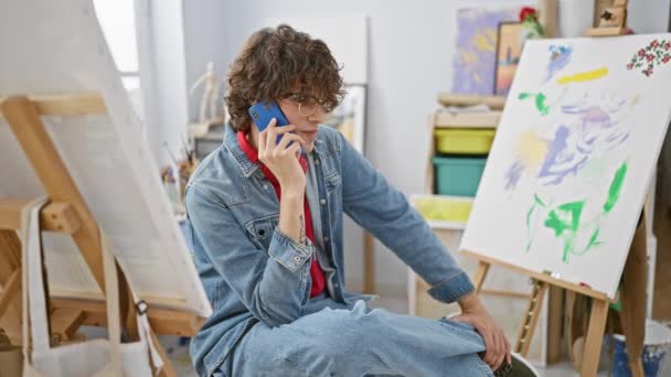 一个卷曲头发 头戴粗斜纹棉布的男人在一间明亮的画室里用帆布和颜料进行电话交谈 — 图库视频影像