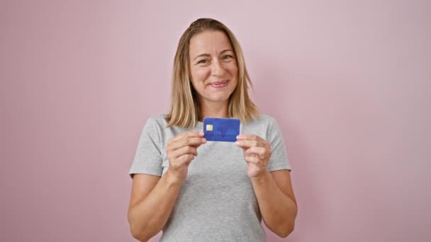 快乐的金发女青年自信地拿着信用卡 在孤立的粉色背景下放弃了大拇指的姿势 — 图库视频影像