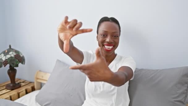 快乐的非洲裔美国女人躺在卧室里 躺在床上 用富有创意的手势美丽地塑造着她的笑脸 传达着摄影的理念 — 图库视频影像