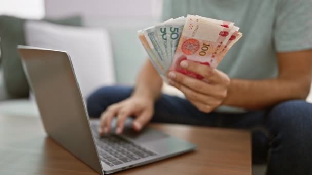 年轻人坐在家里的沙发上 一边拿着人民币钞票 一边埋头于笔记本电脑上的网上金融 — 图库视频影像