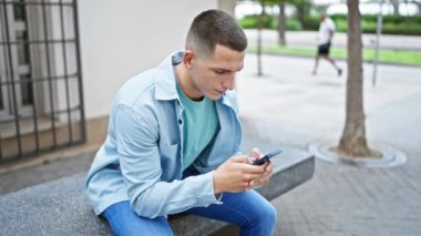 Genç İspanyol adam şehir merkezinde bankta otururken akıllı telefon kullanıyor..