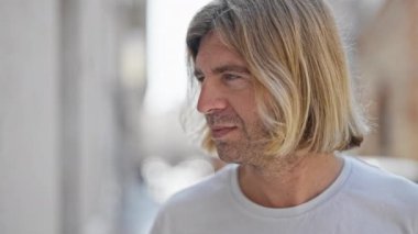 Güler yüzlü sarışın bir adamın samimi bir portresi. Uzun saçlı, güneşli bir sokak ortamında dikiliyor..