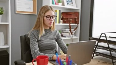 Modern bir ofiste masasında çalışan gözlüklü, şaşırmış sarışın bir kadın..