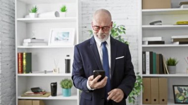 Takım elbiseli olgun bir işadamı akıllı telefon kullanıyor ve modern bir ofis ortamında başparmak kaldırıyor.