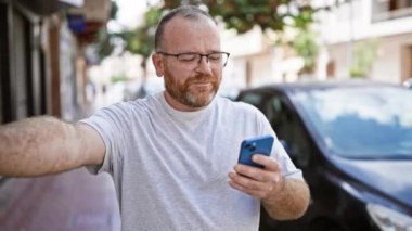 Neşeli, gözlüklü, sakallı, orta yaşlı, beyaz bir adam canlı, şehir sokağında telefonundan bangır bangır müzik dinliyor