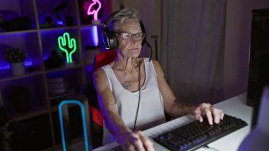 Kendine güvenen, gri saçlı, son sınıf öğrencisi, gece oyun odasında fütüristik video oyunu oynarken gülümseyen ve modern teknolojiyle eğlenen bir kadın.