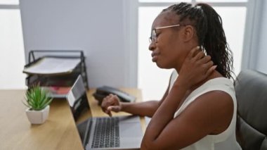Kıvırcık saçlı yetişkin bir kadın modern bir ofis içinde dizüstü bilgisayarla çalışırken boyun ağrısı çekiyor..