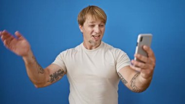 Sarışın bir adam, mavi bir arkaplana karşı akıllı telefonuyla selfie çekiyor ve çeşitli mutlu ifadeler sergiliyor..