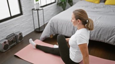 Çekici, genç, sarışın bir kadın yatak odasında uzanırken gülümsüyor ve sabah sporu için ısınıyor.