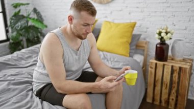 Yakışıklı bir adam modern bir yatak odasında rahat bir yatakta sarı bir bardağın yanında akıllı telefon kullanıyor..