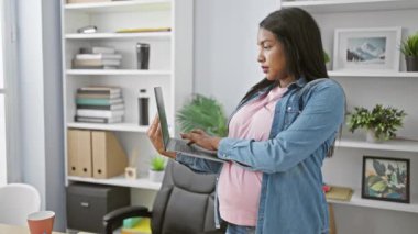 Gülümseyen hamile iş kadını karnına dokunuyor, ofiste dizüstü bilgisayarla çalışıyor, özgüven ve başarı sergiliyor.