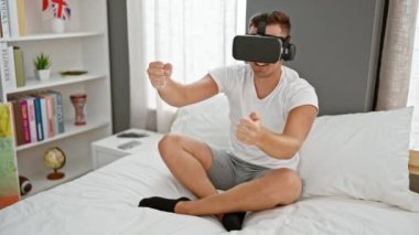 VR kulaklıklı genç adam modern bir yatak odasında heyecanla el kol hareketi yapıyor.
