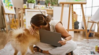 Gülümseyen genç İspanyol kadın sanatçı sevgili köpeğini öpüyor, stüdyo zemininde oturuyor, sanatını dizüstü bilgisayarla icra ediyor, öğrenme sürecinde neşe yaratıyor..