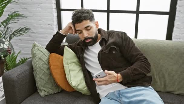 一个留着胡子的沉思的男人坐在屋里的沙发上 在时尚的客厅里拿着电话 — 图库视频影像