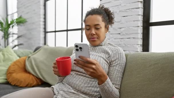 在舒适的客厅里 一个头发卷曲的女人坐在沙发上 手里拿着一个红色的杯子和智能手机 这让她很吃惊 — 图库视频影像