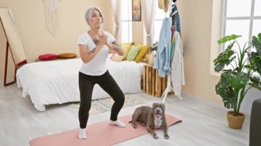 Orta yaşlı, kır saçlı kadın yatak odasında bacaklarını çalıştırırken köpeğini eğitiyor. Formda ve sağlıklı kalmak için bir yaşam tarzı seçimi.