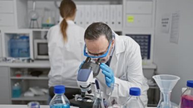 Birlikte çalışarak, laboratuvarda oturan iki ortak, kollarını kavuşturmuş, mikroskobun arkasında, yeni tıp araştırmaları keşfederken gülümsüyorlar..