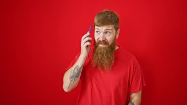 Aşırı neşeli genç kızıl saçlı adam, izole edilmiş bir kırmızı arka planda telefonla konuşurken kendinden geçmiş bir şekilde kutlama yapıyor.