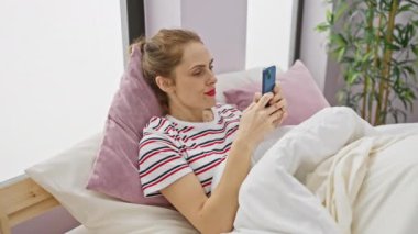 Çizgili tişörtlü rahat bir kadın rahat bir yatak odası ortamında akıllı telefonunu kullanmayı sever..