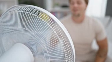 Sakallı sarışın bir adam modern bir yatak odasında elektrikli vantilatörün yanında dinleniyor.