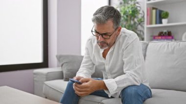 Evdeki kanepede oturan beyaz saçlı İspanyol genç adam akıllı telefon kullanıyor.