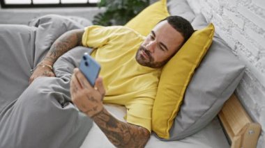 Dövmeleri olan yakışıklı bir adam. Akıllı telefon kullanarak yatakta uzanıyor. Yatak odasının içinde beyaz tuğlalar var..