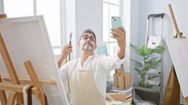 Gülümseyen genç, gri saçlı İspanyol ressam resim stüdyosunda fırçalar ve tuvaller arasında akıllı telefonuyla neşeli bir selfie çekiyor.