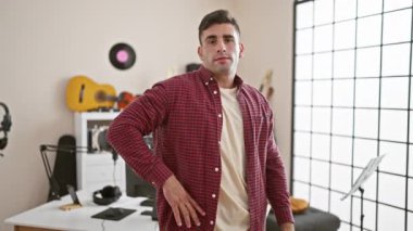 Neşeli bir tebessümle duran, kollarını müzik stüdyosunda kavuşturan, modern bir müzisyenin somut örneği olan kendinden emin, genç İspanyol bir adam.